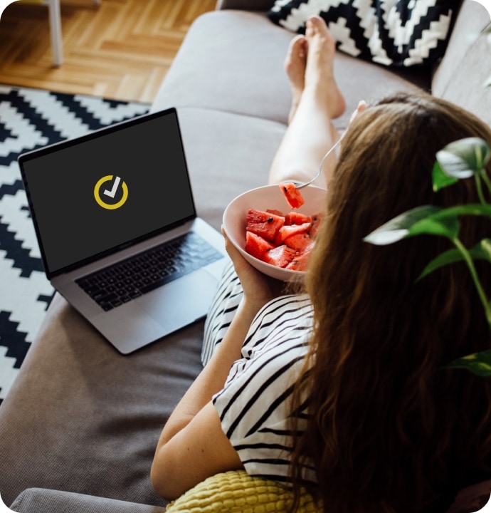 Vrouw liggend op een bank die fruit eet met naast haar een laptop met het Norton-logo op het scherm